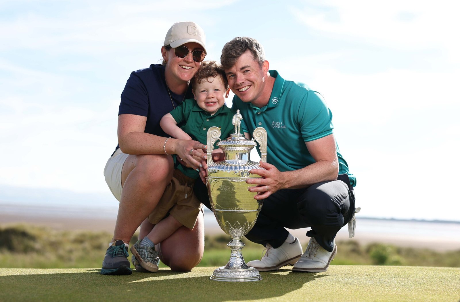 A smiling family of three holds a large silver golf trophy on a sunny course, posing for a celebratory photo.