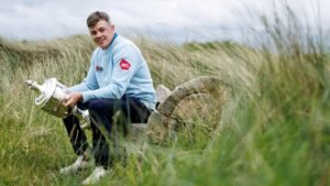 Man in a pale blue sweatshirt sits on a log in tall grass, proudly holding a large silver trophy and smiling outdoors.