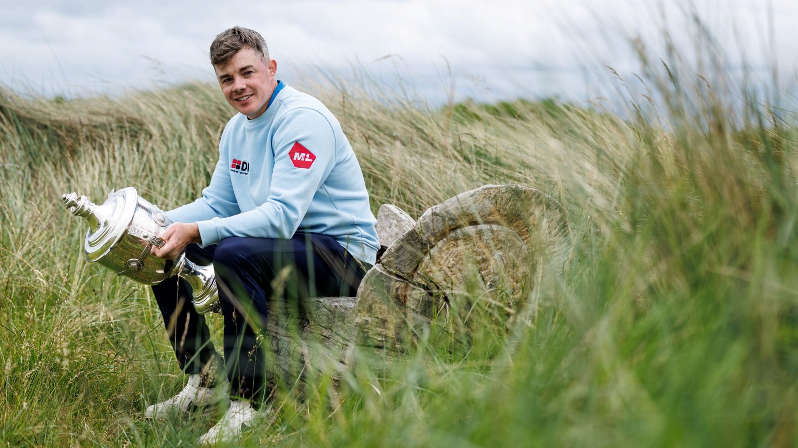 Stuart Grehan - Talk Golf Ireland Man in a pale blue sweatshirt sits on a log in tall grass, proudly holding a large silver trophy and smiling outdoors.