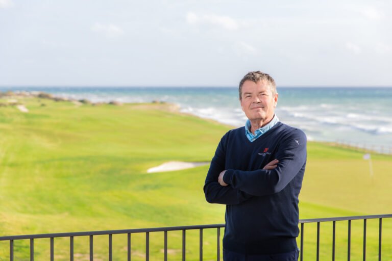 Man in a navy sweater stands with arms crossed near a railing, ocean and grassy park in the background.