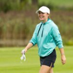 Smiling woman in a turquoise hoodie and white cap walking on a golf course, holding a golf glove.