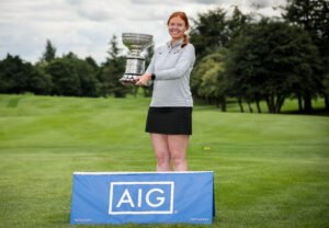 - Talk Golf Ireland Female golfer on a golf course proudly holding a large silver trophy, with a blue AIG sign in the foreground.