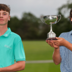 Two young golfers pose outdoors, each holding a trophy after a win on a golf course.