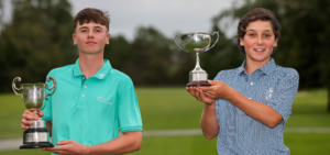 Two young golfers pose outdoors, each holding a trophy after a win on a golf course.
