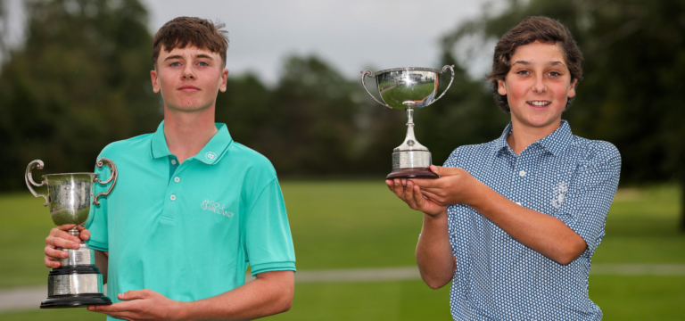 Two young golfers pose outdoors, each holding a trophy after a win on a golf course.