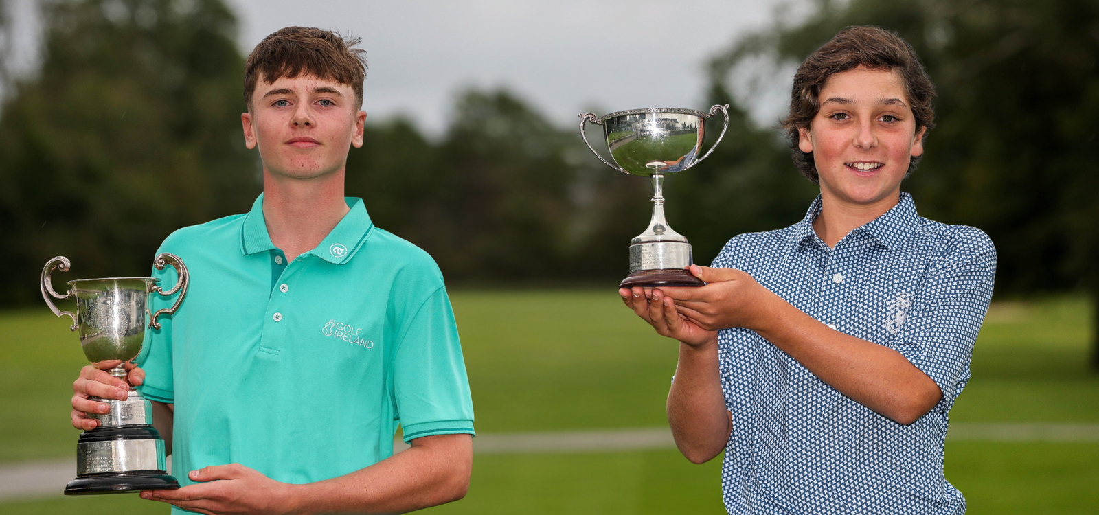 Two young golfers pose outdoors, each holding a trophy after a win on a golf course.