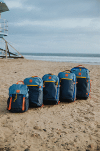 AlphaPacks-PacificColorway 002 - Talk Golf Ireland Blue backpacks lined up on a sandy beach with the ocean in the background and a lifeguard tower to the left.