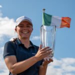 Smiling woman in a navy polo and white cap holds a tall glass trophy; an Irish flag waves in the blue sky behind her.