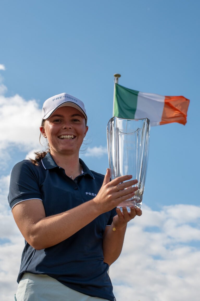 Smiling woman in a navy polo and white cap holds a tall glass trophy; an Irish flag waves in the blue sky behind her.