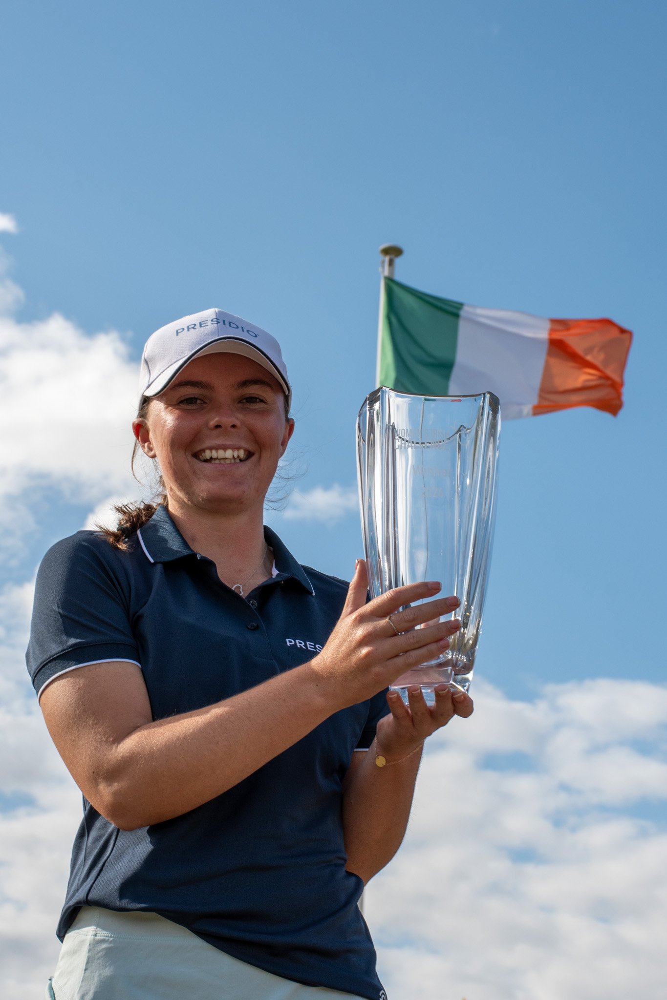 Smiling woman in a navy polo and white cap holds a tall glass trophy; an Irish flag waves in the blue sky behind her.