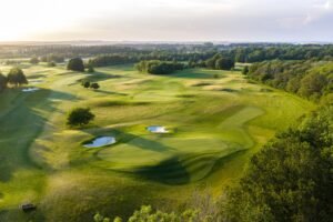 Aerial view of a sprawling golf course with greens, fairways, sand bunkers, and surrounding trees under soft sunlight.