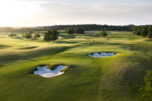Basingstoke 4th green credit James Hogg 002 - Talk Golf Ireland Wide view of a sunlit golf course with rolling greens, sand bunkers, and clusters of trees at golden hour