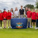 Group of young golfers and coaches posing with trophies on a table at a sunny golf course.