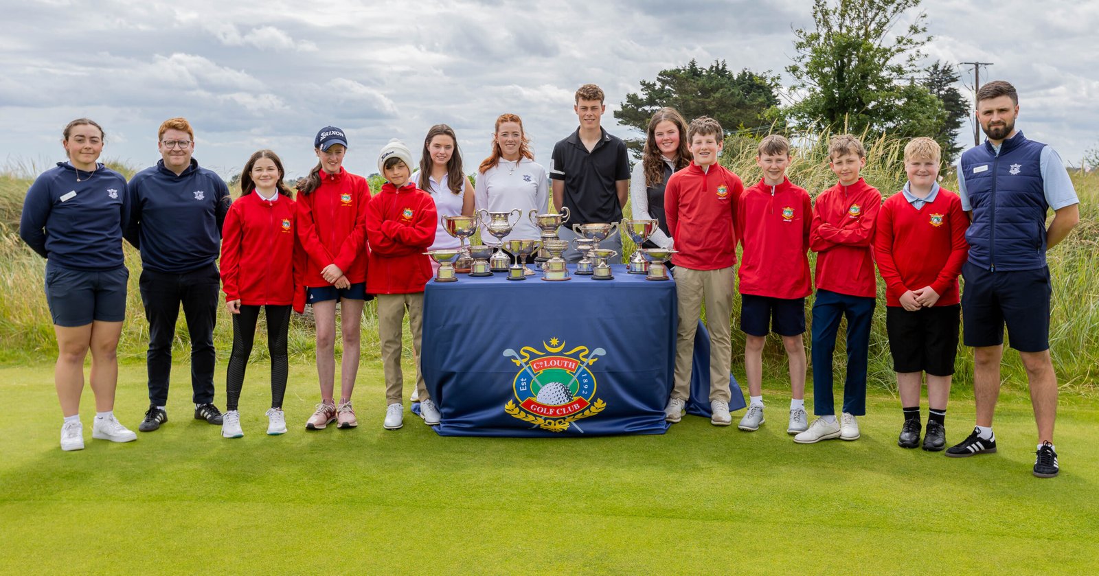 Group of young golfers and coaches posing with trophies on a table at a sunny golf course.