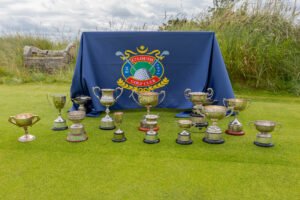 - Talk Golf Ireland Row of silver golf trophies arranged on a green lawn in front of a blue-draped table with a club crest banner behind them