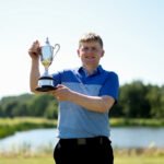 Young man in a blue polo holding a silver trophy outdoors on a sunny day near a park or golf course setting