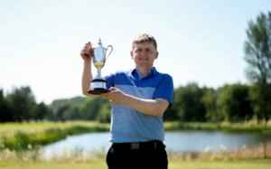 Young man in a blue polo holding a silver trophy outdoors on a sunny day near a park or golf course setting