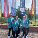 Five people in teal blazers pose together outdoors, with tall colorful flags behind them on a sunny day.