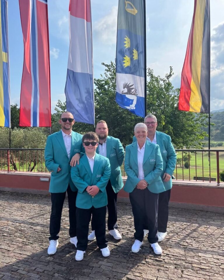 Five people in teal blazers pose together outdoors, with tall colorful flags behind them on a sunny day.