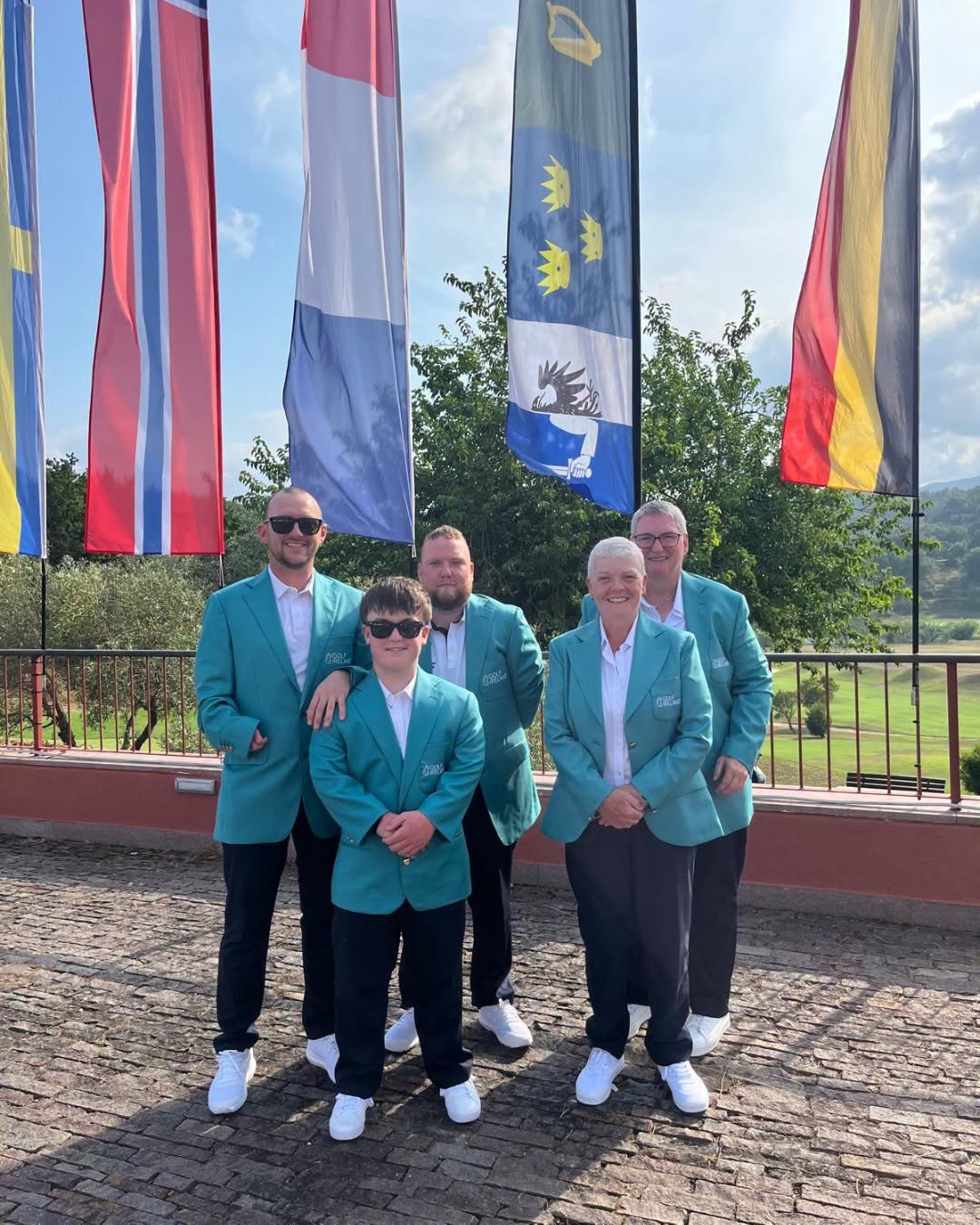 Five people in teal blazers pose together outdoors, with tall colorful flags behind them on a sunny day.