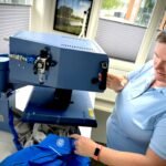 Lab technician wearing a light blue shirt uses a large blue scientific instrument with controls; she is seated at a workstation in a bright room with blinds on the windows.