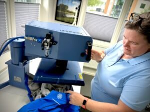 Lab technician wearing a light blue shirt uses a large blue scientific instrument with controls; she is seated at a workstation in a bright room with blinds on the windows.