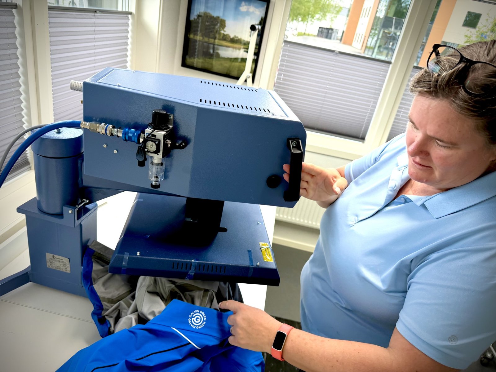 Lab technician wearing a light blue shirt uses a large blue scientific instrument with controls; she is seated at a workstation in a bright room with blinds on the windows.