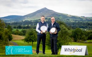 Two men stand outdoors on a golf course, holding large trophies with a scenic mountain backdrop and sponsor banners nearby.