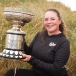 Young woman in a black hoodie sits in tall grass, proudly holding a large ornate silver trophy outdoors.