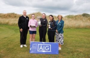 Katie Poots 2 - Talk Golf Ireland Five people posing with trophies on a golf course, with a blue AIG banner in front under a cloudy sky.