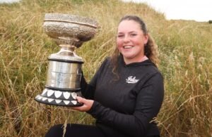 Young woman in a black hoodie sits in tall grass, proudly holding a large ornate silver trophy outdoors.