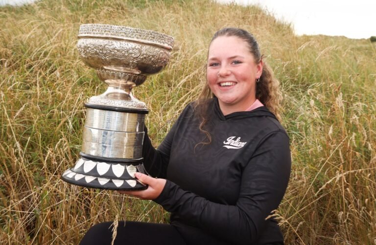 Young woman in a black hoodie sits in tall grass, proudly holding a large ornate silver trophy outdoors.
