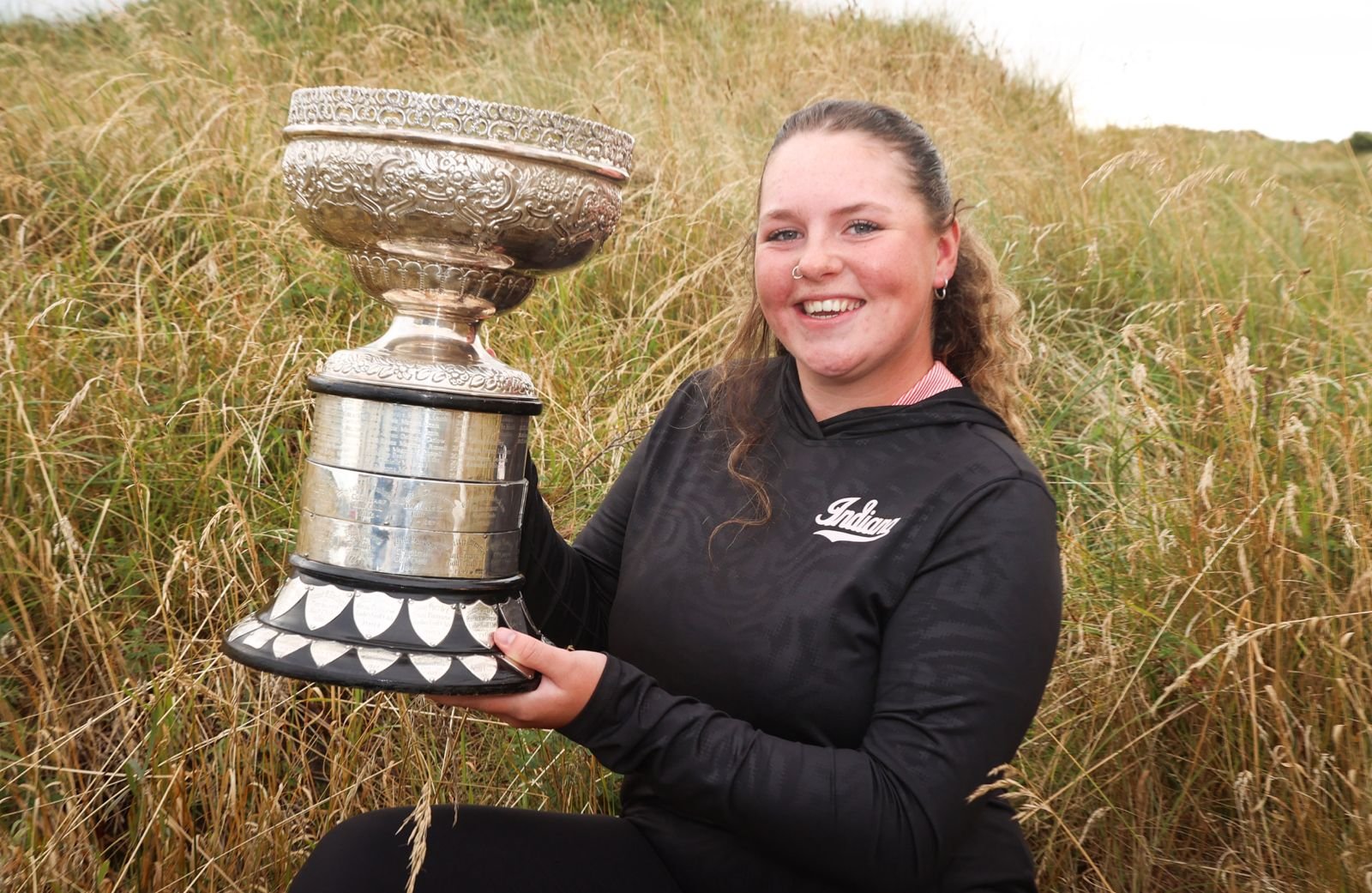 Young woman in a black hoodie sits in tall grass, proudly holding a large ornate silver trophy outdoors.
