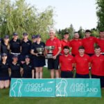 Group of golfers posing outdoors with trophies; team in navy and red shirts beside a 'Golf Ireland' banner.