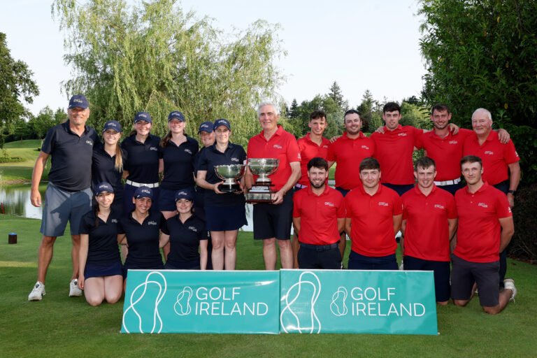 Group of golfers posing outdoors with trophies; team in navy and red shirts beside a 'Golf Ireland' banner.