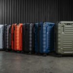 Row of colorful hard-shell suitcases on wheels lined up against a dark vertical-panel wall in a studio setting