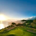 Sunlit coastal golf course at sunset with green fairways and distant hills by the sea.