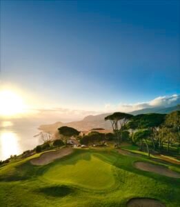 Sunlit coastal golf course at sunset with green fairways and distant hills by the sea.
