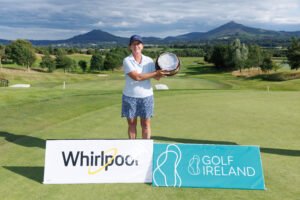 RDR_4013 - Talk Golf Ireland Woman on a golf course holding several round trophies or plaques, smiling, with hills and mountains in the background; banners advertising Whirlpool and Golf Ireland in the foreground.