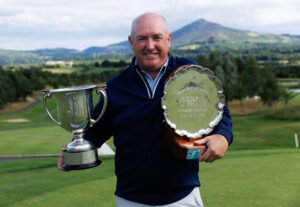 RDR_4160 - Talk Golf Ireland Smiling man in a navy jacket holds a large silver trophy and a round shield plaque outdoors on a golf course with hills in the background.