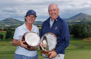 Two smiling golfers pose with large silver trophies on a sunny golf course with hills in the background.