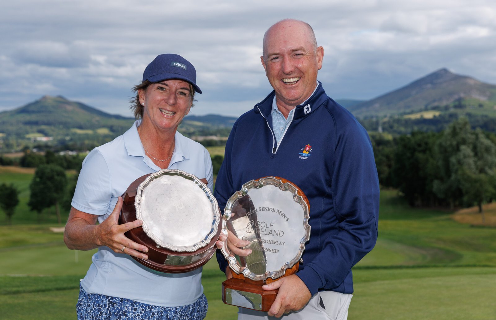 Two smiling golfers pose with large silver trophies on a sunny golf course with hills in the background.