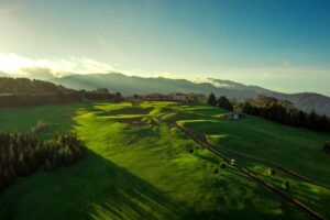 Santo da Serra Golf010Digital Couple small 002 - Talk Golf Ireland Aerial view of a green golf course with a red-roofed clubhouse, winding cart paths, and distant hills at sunset