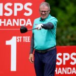 Golfer in a teal shirt mid-swing with a golf club, against a red ISPS backdrop.