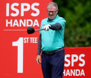 Golfer in a teal shirt mid-swing with a golf club, against a red ISPS backdrop.