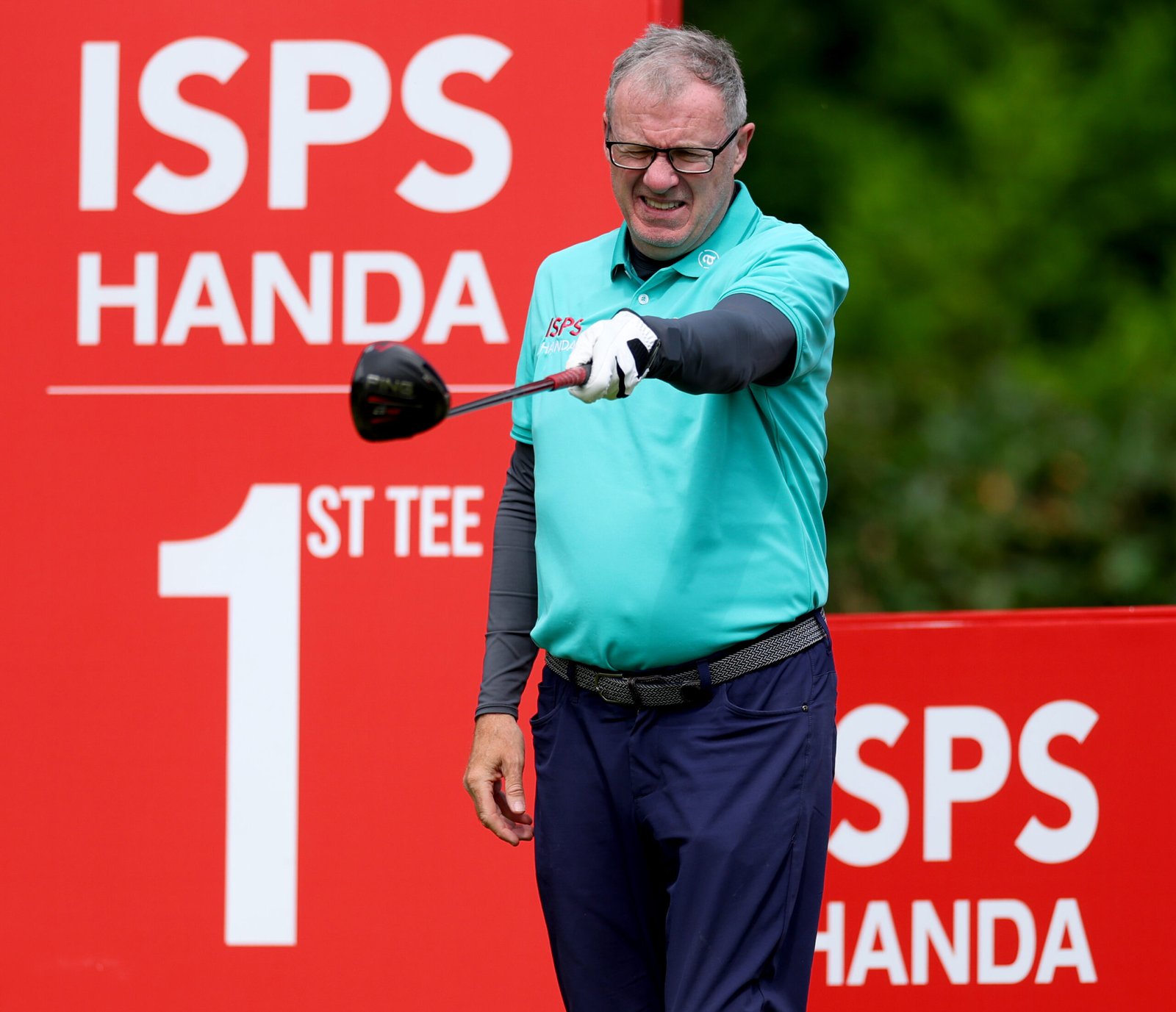 Golfer in a teal shirt mid-swing with a golf club, against a red ISPS backdrop.