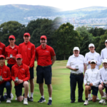 Group photo of a golf club team on a fairway, left in red uniforms and right in white uniforms, each member holding medals.