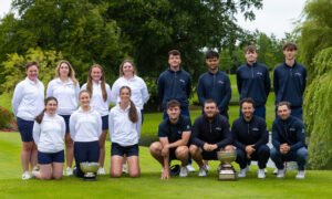 Group of golfers posing outdoors on a well-manicured lawn with two trophies in front, wearing white and navy uniforms.