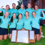 Ten teammates in matching turquoise tops pose on a lawn, arms raised in celebration, with a wooden trophy board on the grass in front.
