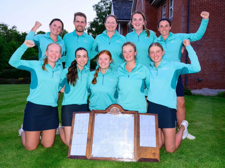 Ten teammates in matching turquoise tops pose on a lawn, arms raised in celebration, with a wooden trophy board on the grass in front.